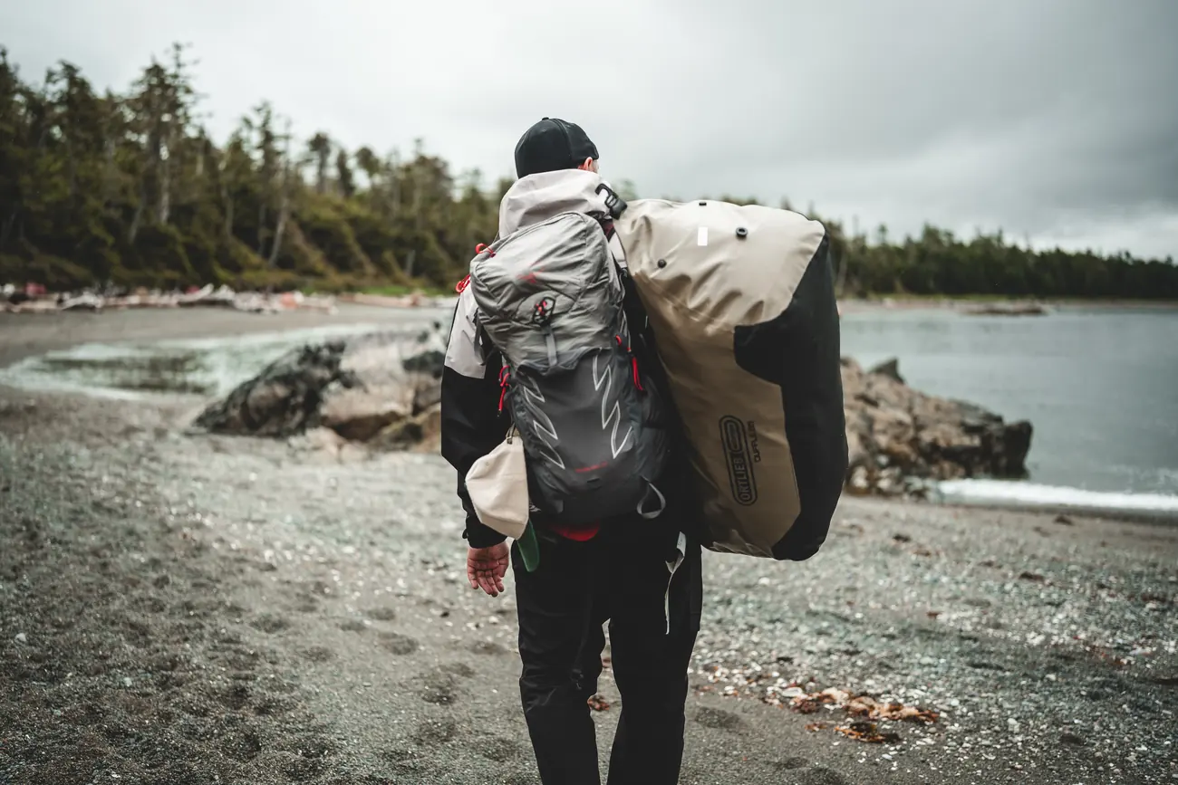 Person mit großem Expeditionsrucksack am Kiesstrand vor dem Küstenregenwald von Porcher Island